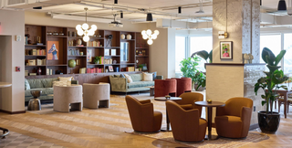 A sophisticated coworking lounge featuring plush seating, elegant lighting fixtures, and a wall of bookshelves filled with books and decorative items. The space includes comfortable sofas, armchairs, and small round tables, creating an inviting atmosphere for work or relaxation. Large windows and indoor plants enhance the bright and airy feel of the room.
