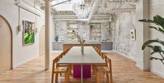 A modern, loft-style dining space featuring a long table with wooden chairs, situated under a chandelier with circular light fixtures. The room boasts a white, exposed brick wall, wooden floors, and a combination of open shelving and cabinetry along the back wall. Artwork adorns the walls, adding a touch of sophistication. The space is bathed in natural light from large windows or skylights above, and a potted plant adds a hint of greenery to the right side.